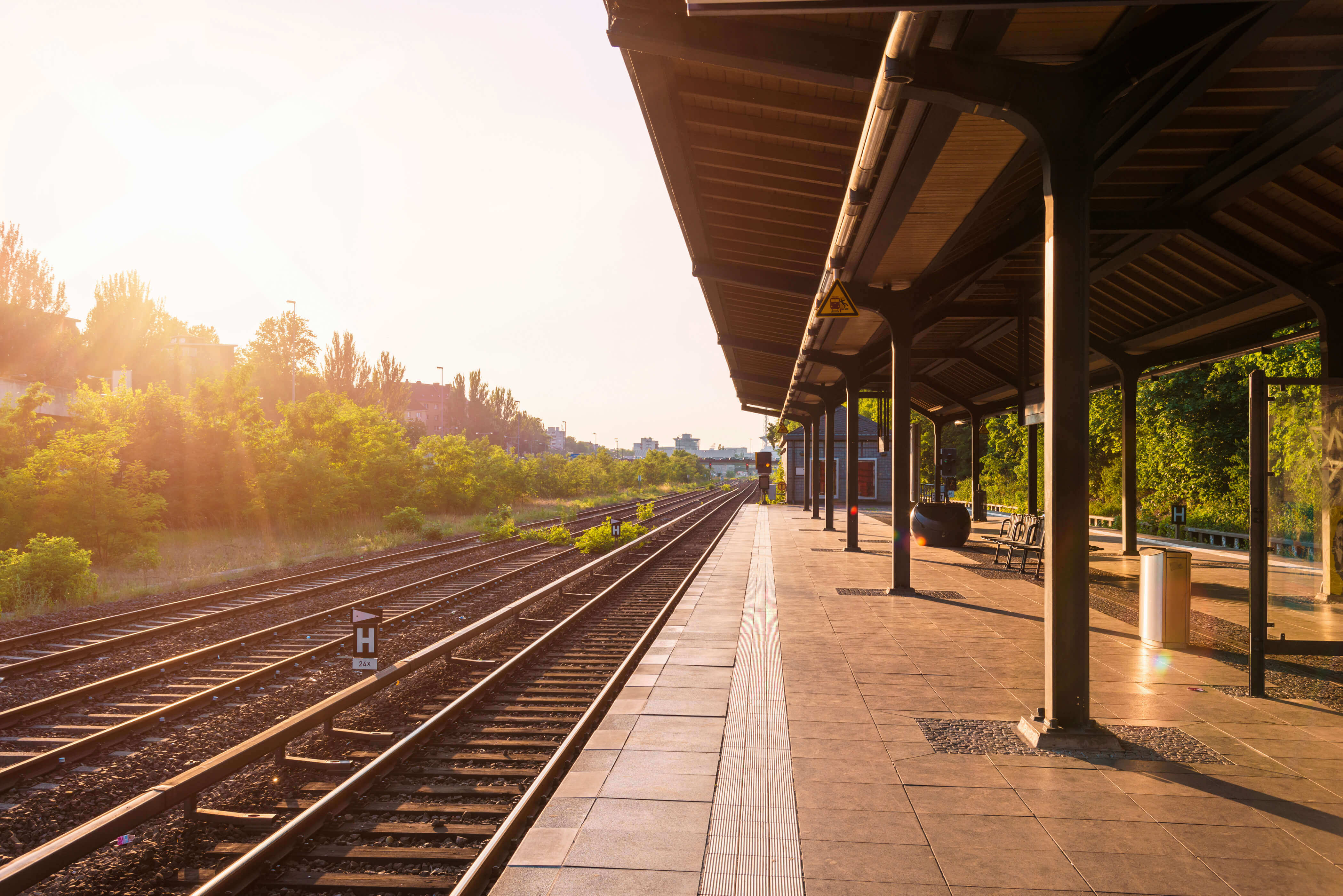 A train platform alongside rail tracks, surrounded by trees in the sunshine